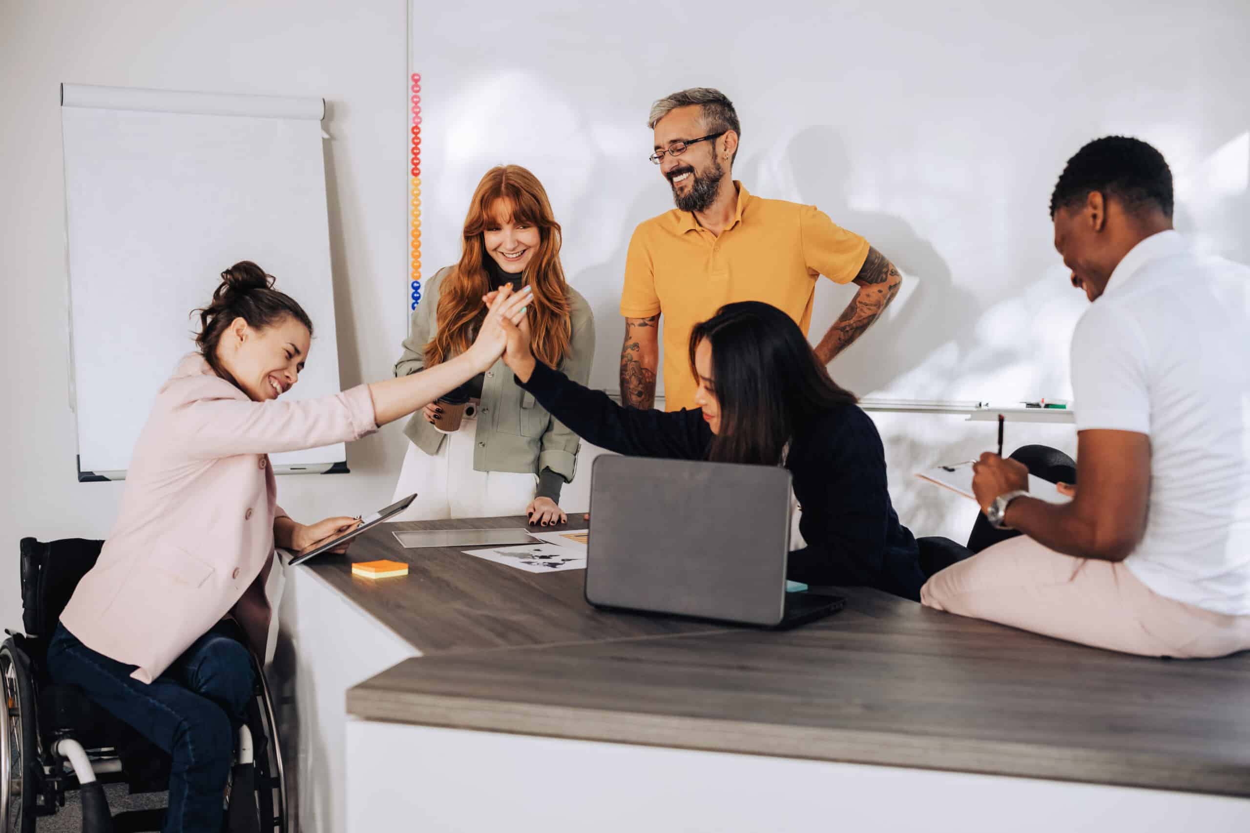 A multicultural boss is congratulating a businesswoman who uses a wheelchair success on a project while sitting with her team at office. Achievement and success on a project, teamwork and cooperation.